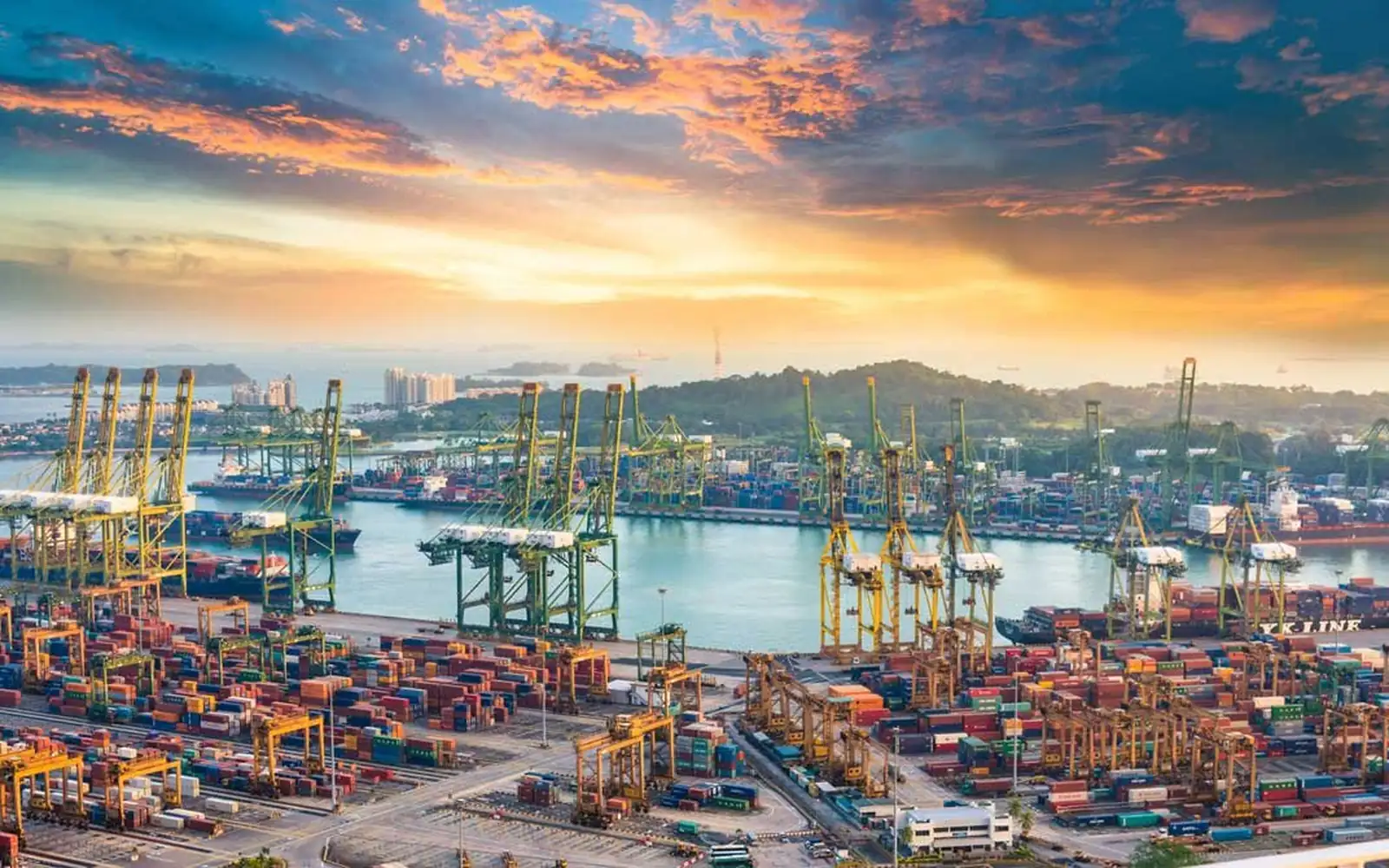 Aerial view of a container terminal at sunset, quay cranes lining the wharf and stacked containers behind them.